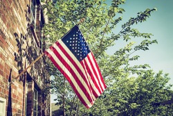 Patriotic American flag in front of a brick home, trees and blue sky background. Vintage filter effects.