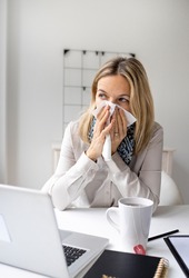 Sick business woman in office feeling unwell sitting in front of her laptop blowing her nose, stressed femal employee have anxiety attack at workplace