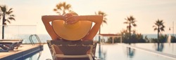 Rear view woman wear hat lying on deckchair near pool, put hands behind head relaxing, take sun bath, sea palm tree empty swimming pool scenery on background. Summer holidays, vacation, travel concept