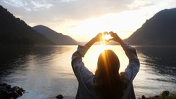 Close up on female's hands making a heart shape finger frame on lake forest landscape during sunset. People love nature environment concept