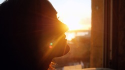 Woman looking standing near the window with view on sunset in city with lense flare effects