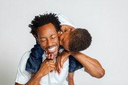An African American boy on the shoulders of his father, leans over to kiss him on the cheek.  His father laughs with closed eyes.