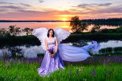 young girl with angel wings and flying veil sits on a meadow with bouquet