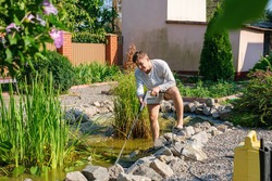 Young smiling caucasian man cleans artificial garden pond bottom with high-pressure washer nozzle from mud and sludge. Spring and summer pond care work