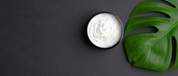 Jar with cosmetic cream and tropical monstera leaf on black background. Natural organic beauty product for body, hand, face skin treatment. Flat lay, top view. Skincare concept.
