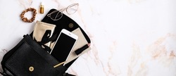 Black leather women's bag with smartphone, pouch, paper notebook, lipstick, feminine glasses, scrunchie on marble table. Beauty blog banner design. Flat lay, top view, copy space.