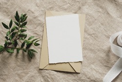 Blank paper card mockup, envelope, green branch, silk ribbon on linen tablecloth. Wedding invitation card design, greeting card template.