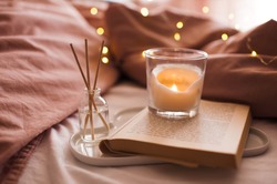 Burning candle with aroma sticks in bottle on tray with open book in bed over glowing Christmas lights close up. Cozy atmosphere at home. Good morning. Selective focus. 