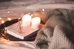 Aroma sticks with liquid scented water in bottle with burning candles staying on open book and knitted textile in bed closeup over lights at background . Winter holiday season. 