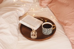 Cup of black fresh coffee with liquid aroma perfume in glass bottle with wood sticks and open paper book in tray in bed with pink blanket and duvet. Cozy home atmosphere. Aromatherapy. 