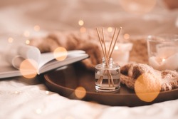 Home perfume in glass bottle with wood sticks, scented burn candles, open paper book and knit wool textile on ray in bedroom close up. Aromatherapy cozy atmosphere lifestyle. Winter warm xmas season. 