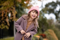 Happy cute child girl 4-5 year old wear knit wool hat and jacket over nature backggound outdoors. Autumn season. Looking at camera. Happiness. Laughing little kid having fun in park. Childhood. 