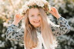 Smiling child girl wear floral wreath and dress posing over blooming tree outdoor at nature background. Looking at camera. Childhood. Springtime. 