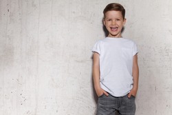 Cute little boy in white T-shirt posing in front of grunge concrete wall. Portrait of fashionable male child. Smiling boy posing, gray wall on background. Concept of children style and fashion.