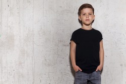 Cute little boy in black T-shirt posing in front of grunge concrete wall. Portrait of fashionable male child, copy space. Boy looks at camera, gray wall on background.