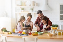 Happy family cooking together on kitchen. Mother and daughter reading recipe to father and son. Dad and boy chopping green vegetable leaf for salad. Home recreation and food preparation on weekend