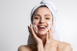Happy laughing young caucasian woman wrapped in head towel washing face with foam soap cosmetic product. Everyday morning hygiene routine. Skincare and wellness, beauty and cosmetology. Portrait