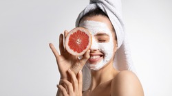 Close up of happy young playful teen girl in moisturizing mask and towel holds grapefruit covers eye. Advertising poster of facial eco-friendly skincare products. Morning beauty routine