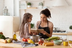 Beautiful young mother reading to adorable daughter on kitchen. Mom and child learning recipe for cooking dinner. Happy family engaged in food preparation at home. Girl sit on table full of vegetable