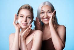 Happy female generations young girl daughter, mature gray-haired woman mother care of facial skin, applying cleansing foam. Women having fun washing faces on blue background. Skincare routine and age