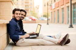 Two friends sitting on the street putting the laptop on the leg and looking with a smile.