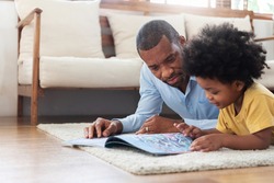 Happy time of African family. African-American Father lying reading book on the floor with his son before traveling to work at living room. Parenthood and childhood concept.