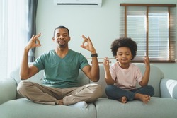 Smiling Black African american father and little son sitting on sofa doing yoga in meditation at home together.