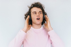 Amazed handsome stylish young male with curly hair wearing pink clothes, with headphones on head, listening favorite music, isolated on white studio background. Copy space for advertisement text