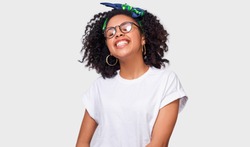 Beautiful dreamy dark-skinned young woman dressed in white t-shirt, feeling happy and enjoying the weather. African American female smiling broadly, wearing round eyewear posing over white studio wall