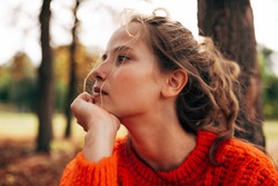 Closeup portrait of a pensive woman looking away, wearing an orange knitted sweater posing on a fall nature background. The beautiful female has a thoughtful expression, resting outdoor in the park.