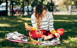 Horizontal image of a student female sitting on the green grass at the college campus on a sunny day, have lunch and studying. The hungry young woman takes a rest learning and eating in the park.