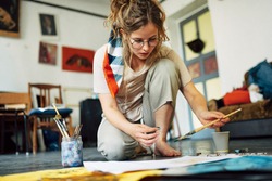 Horizontal image of a pretty female artist sitting on the floor in the art studio and painting on paper with a brush. A woman painter with glasses painting with watercolors in the workshop.