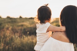 Cropped rear view of Caucasian brunette mother and child daughter embrace each other on meadow sunset. Beautiful brunette woman hugs her toddler girl and looking on sunset background. Motherhood.