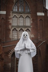 A gloomy portrait of a ghostly pale nun in white robes with black eye sockets and black smudges under them, amid gloomy Gothic architecture. Halloween, horror.