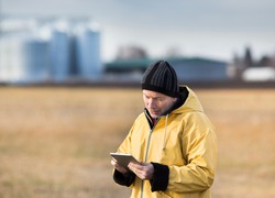 Mature farmer with tablet standing in field in winter time, with grain silos in background
