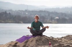 Senior man sitting in lotus position and meditating on sand hill on river coast