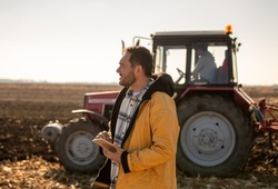 Farmer holding tablet and standing in front of tractor in field in winter time