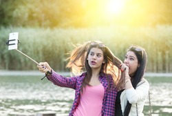 Two pretty teenage girls taking selfie on mobile phone with stick in the nature beside river. Positive and crazy emotions