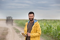 Young farmer with beard holding tablet in front of tractor in field. Seasonal agricultural works
