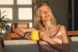 Attractive happy middle aged woman is sitting on  sofa in living room. Smiling adult lady enjoys drinking coffee or tea sitting on couch at home