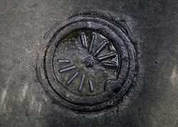 Full frame photograph of a dirty, wet, rusted manhole cover surrounded by dark concrete.