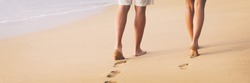 Beach couple walking barefoot on sand at sunset walk honeymoon travel banner - woman and man relaxing together leaving footprints in the sand.