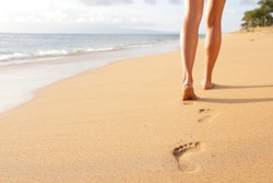 Beach travel - woman walking on sand beach leaving footprints in the sand. Closeup detail of female feet and golden sand on Kaanapali beach, Maui, Hawaii, USA.