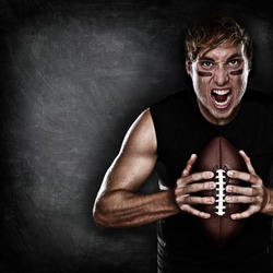 Football player aggressive portrait holding american football on black blackboard background with copy space for text or design. Caucasian male model in his 20s.