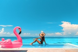 Travel on beach by pool on summer vacation in luxury resort. Woman relaxing in bikini by inflatable pink flamingo toy pool float on ocean turquoise background. Holiday travel destination concept.