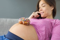Unhealthy food during pregnancy. Sugar temptation craving Asian pregnant woman trying to resist eating a sweet donut on her baby belly. Funny eating cravings concept