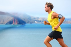 Athlete running man - male runner in San Francisco listening to music on smartphone. Sporty fit young man jogging by San Francisco Bay and Golden Gate Bridge. Jogger training with smart phone armband,