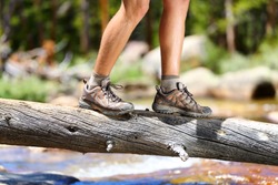 Hiking man crossing river in walking in balance on fallen tree trunk in nature landscape. Closeup of male hiker trekking shoes outdoors in forest balancing on tree. Balance challenge concept.