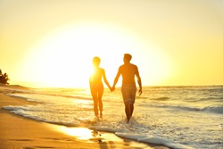 Honeymoon romantic couple in love holding hands walking on beautiful sunset at beach in waterfront. Lovers or newlywed married young couple by the sea enjoying relaxed vacation travel holiday. Hawaii.