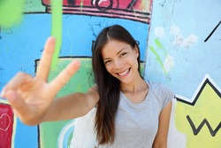 Urban young girl showing v peace sign in city. Cool Asian woman leaning on graffiti background at the Berlin wall, Germany. Modern portrait.